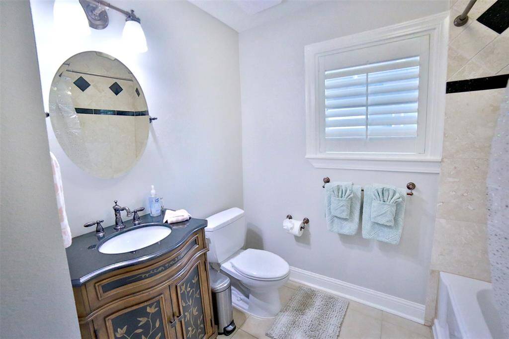 Bright and elegant 3rd bathroom featuring a stylish vanity with a decorative mirror, modern fixtures, and a window with shutters. There's a bathtub, toilet, and neatly arranged towels, creating a fresh and inviting space.