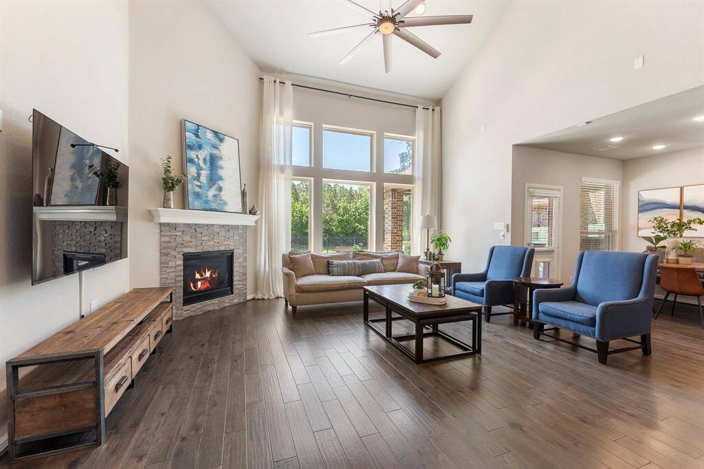 This spacious living room features high ceilings and large windows that provide natural light. It includes a cozy fireplace, modern ceiling fan, and stylish wood flooring. The open layout flows seamlessly for perfect entertaining.