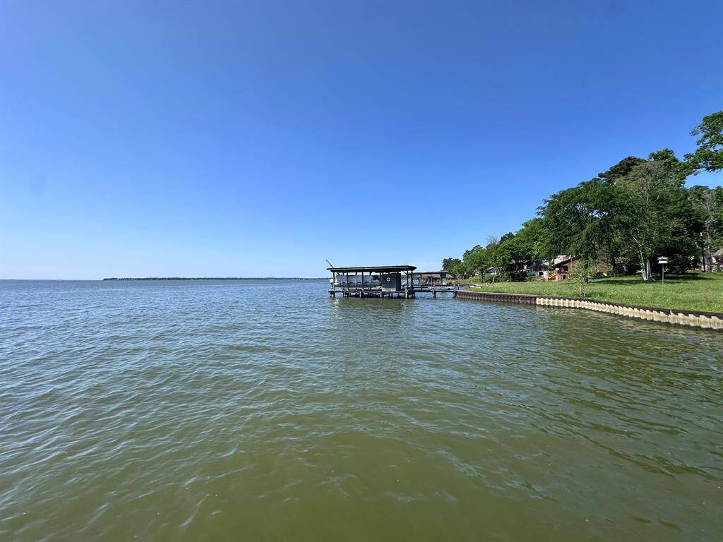 This photo showcases a serene lakeside view with a clear blue sky. There's a dock extending into the water, surrounded by lush greenery and nearby homes. Ideal for those seeking a peaceful waterfront property.