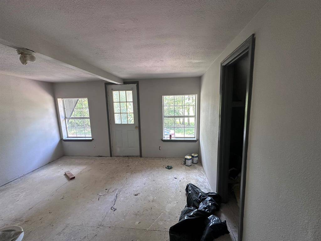 This photo shows a small room with unfinished flooring and two windows providing natural light. There is a door leading outside, and the walls are painted light gray. Some paint cans and a plastic bag are on the floor, suggesting ongoing renovation work.
