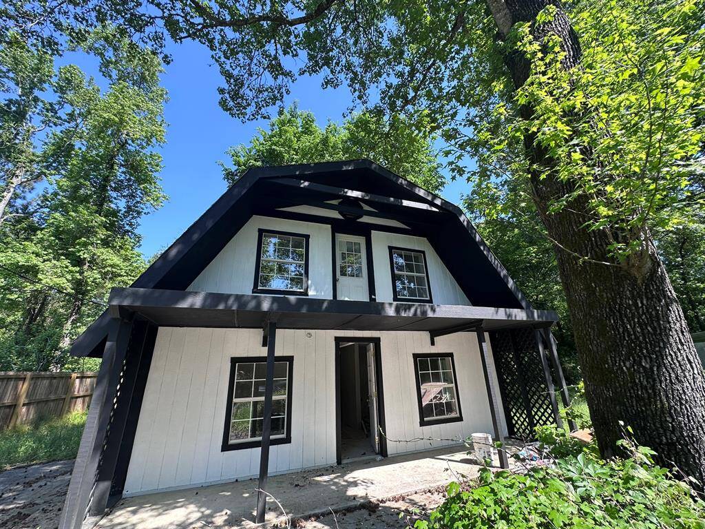 Charming two-story home with a unique roof design, nestled in a wooded area. It features a black and white exterior, large windows, and a cozy porch, offering a serene, natural setting.