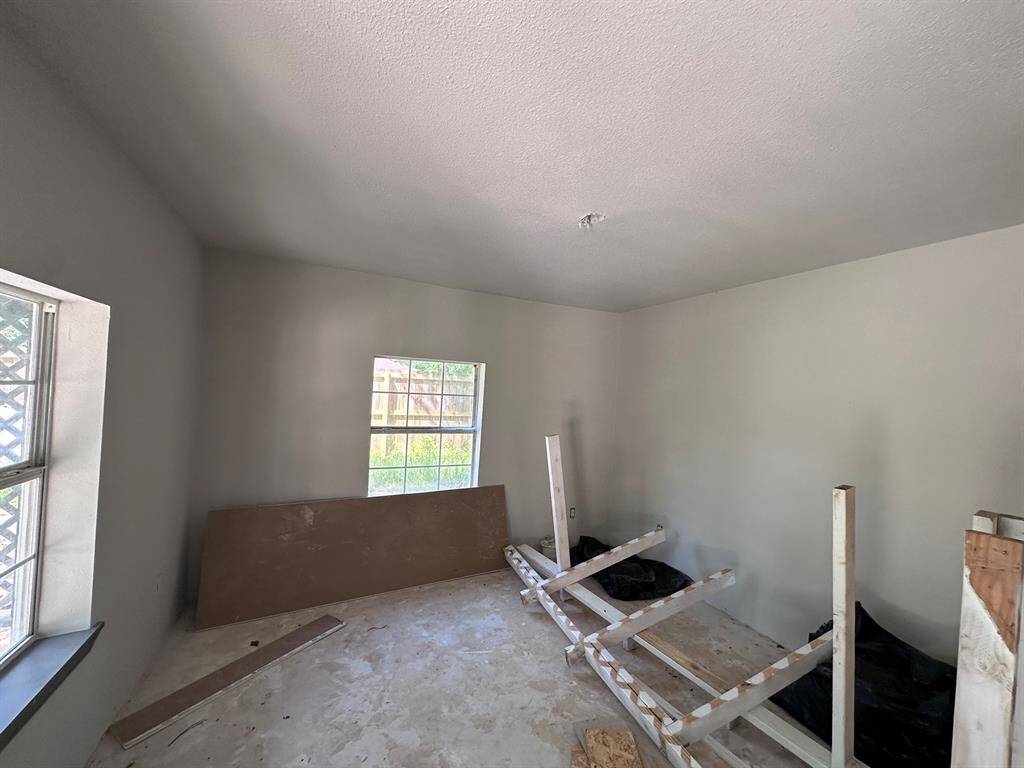 This photo shows an unfinished room with bare walls and a concrete floor. There are two windows allowing natural light, and some construction materials are present. The space appears to need further work and finishing.