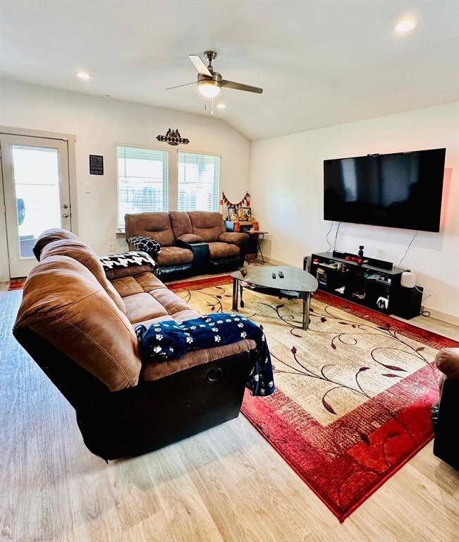 Spacious living room with cozy brown seating, a large wall-mounted TV, and stylish wood flooring. Natural light streams in through multiple windows, and a ceiling fan adds comfort.