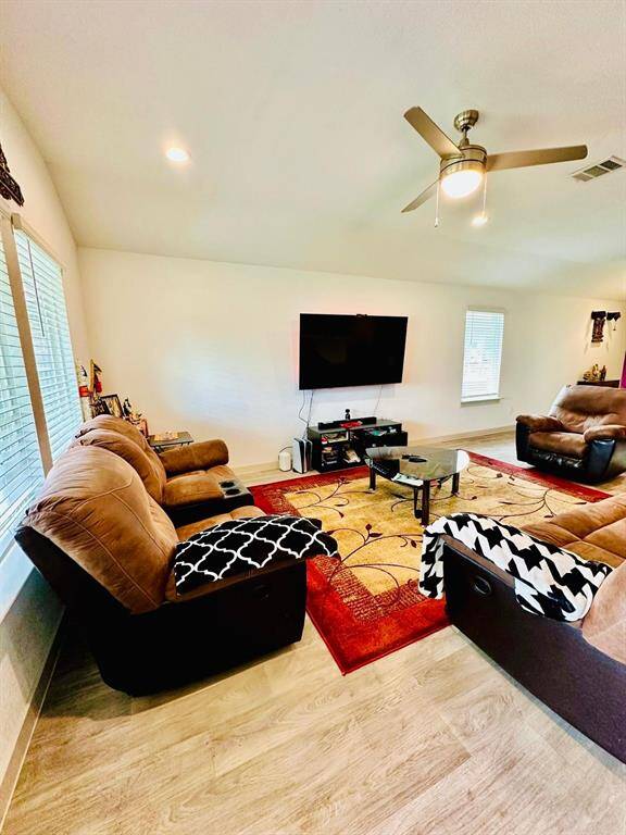 Spacious living room with cozy brown seating, a large wall-mounted TV, and stylish wood flooring. Natural light streams in through multiple windows, and a ceiling fan adds comfort.