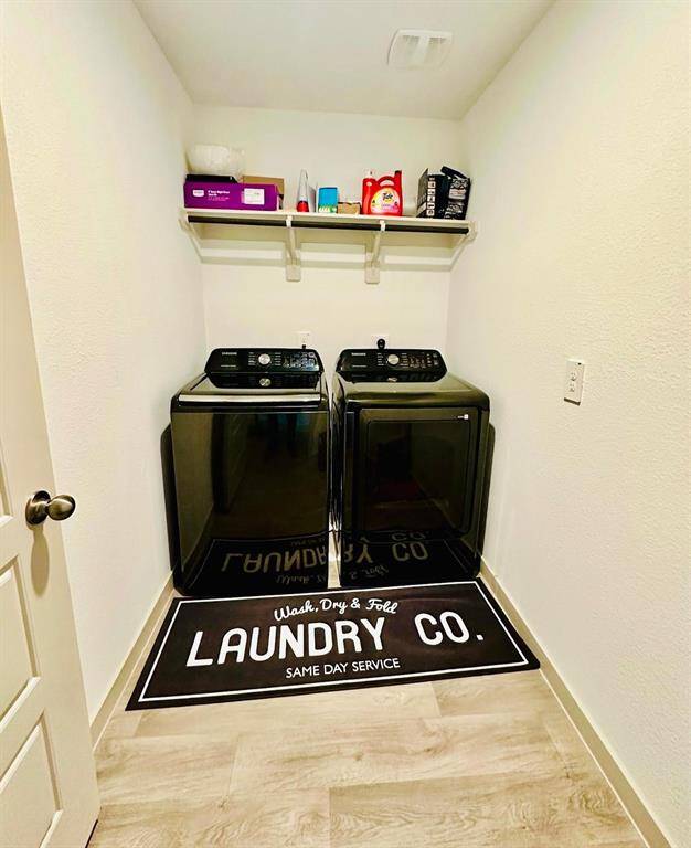 This photo shows a compact laundry room featuring modern black washer and dryer units. Above them, a shelf holds various laundry supplies. The room has clean white walls, light wood flooring, and a decorative mat with a laundry theme.