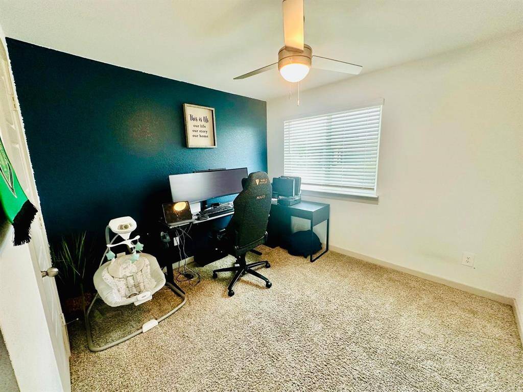 Cozy home office with a modern ceiling fan, plush carpet, and a feature wall painted dark blue. It includes a desk setup with dual monitors and a comfortable chair, plus a window providing natural light.