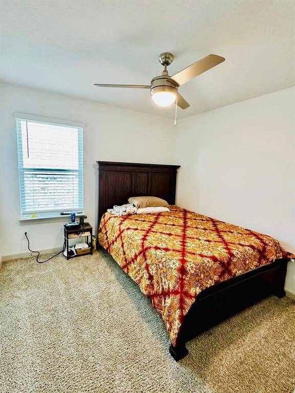 This cozy bedroom features a comfortable bed with a decorative cover, a ceiling fan for airflow, plush carpet, and a window providing natural light. Ideal for relaxation.