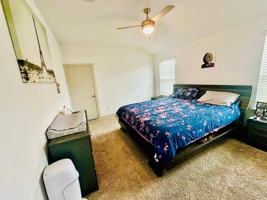 This bedroom features a cozy aesthetic with carpet flooring, a ceiling fan, and ample natural light from two windows. It includes a large bed with a floral comforter, modern furniture, and neutral-colored walls adorned with artwork.