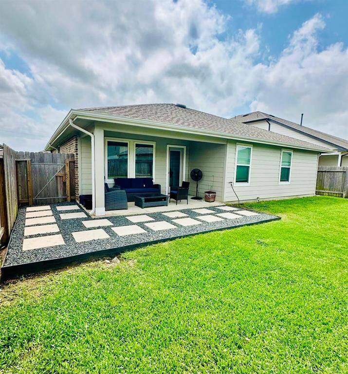 This photo shows the backyard of a home with a covered patio. It features outdoor seating on a checkerboard-style stone area, surrounded by grass, and enclosed by a wooden fence. The space is ideal for relaxing or entertaining.
