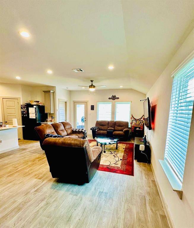 This welcoming living space features an open layout with modern wood flooring, ample natural light from large windows, and cozy seating. A mounted TV and adjacent kitchen area enhance functionality and comfort.