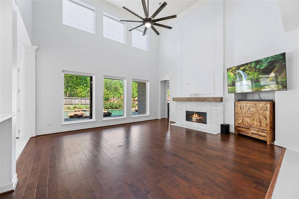Airy and inviting family room with double-height ceilings, a wall of windows overlooking the sprawling pool, a cozy fireplace, and modern fan fixture.