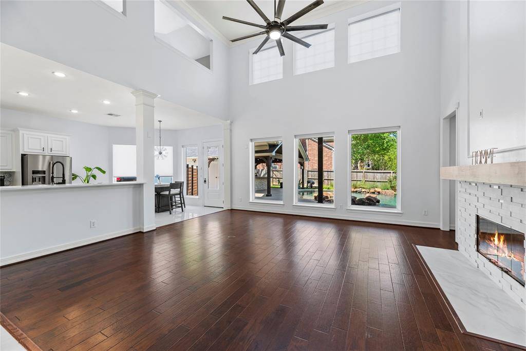 Spacious two-story living room featuring rich wood floors, a sleek stacked stone fireplace, and expansive updated windows offering serene views of the backyard pool.