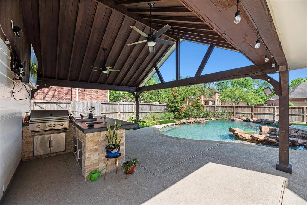 Spacious covered outdoor kitchen and lounge area featuring a built-in stainless-steel Coyote grill, a gas burner, granite counters, TV, and ceiling fans, all overlooking the sparkling pool.