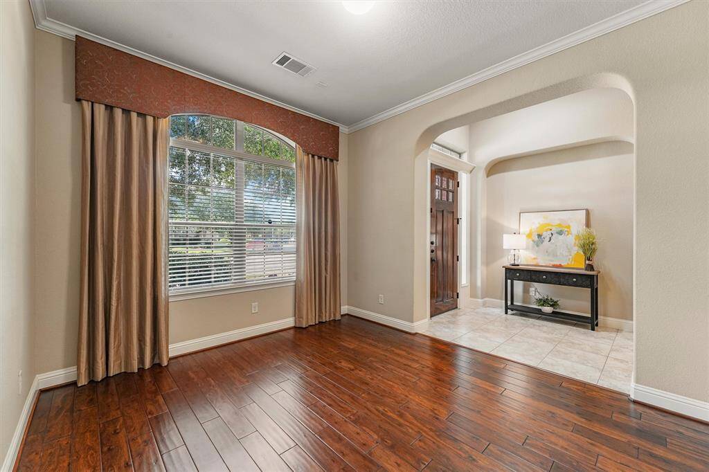 The office features warm wood flooring, a large window with elegant drapery, and an arched doorway leading to a tiled foyer. The space is accented with soft lighting and tasteful decor, creating a welcoming atmosphere.