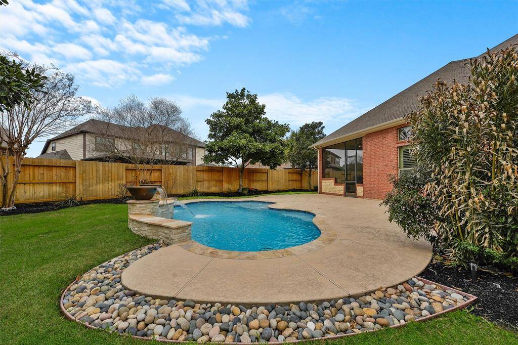 This backyard features a refreshing in-ground pool with a curved design adorned with 3 waterfalls, surrounded by decorative stones which are incorporated into the french drain system to ensure proper drainage throughout the backyard.