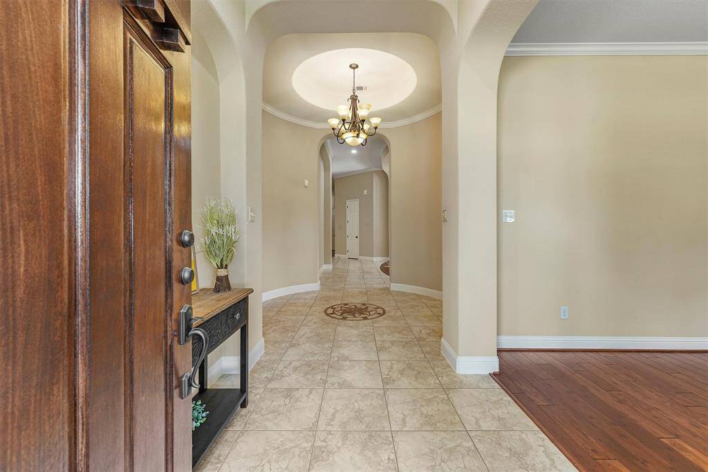 This inviting entryway features a warm wooden 8 ft. door, elegant tiled flooring, and a charming chandelier. The arched hallway leads to a spacious interior with a mix of tile and wood flooring, offering a welcoming and stylish first impression.