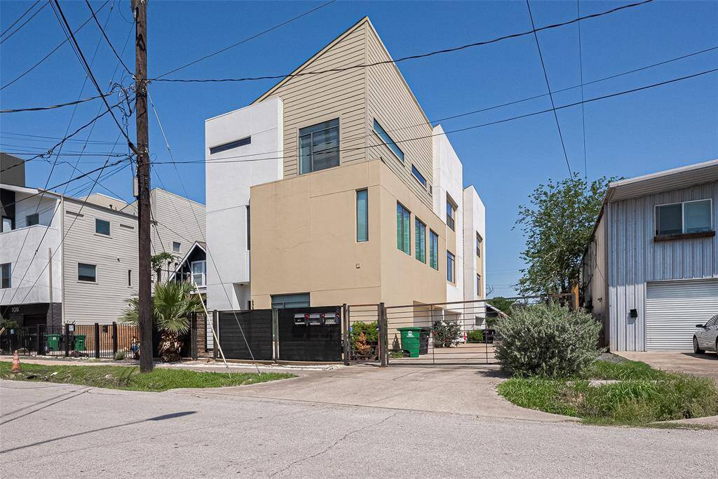 Modern multi-story home with a unique angular design, featuring a mix of beige and white exterior. The property is enclosed by a fence and has a driveway for parking. Located in an urban setting with neighboring contemporary homes.