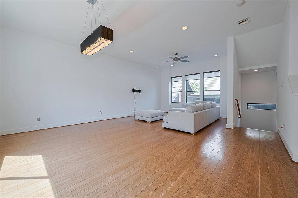 Bright and spacious living area with wood flooring, modern lighting, and large windows. Features a cozy sectional sofa and ceiling fan, perfect for relaxation.