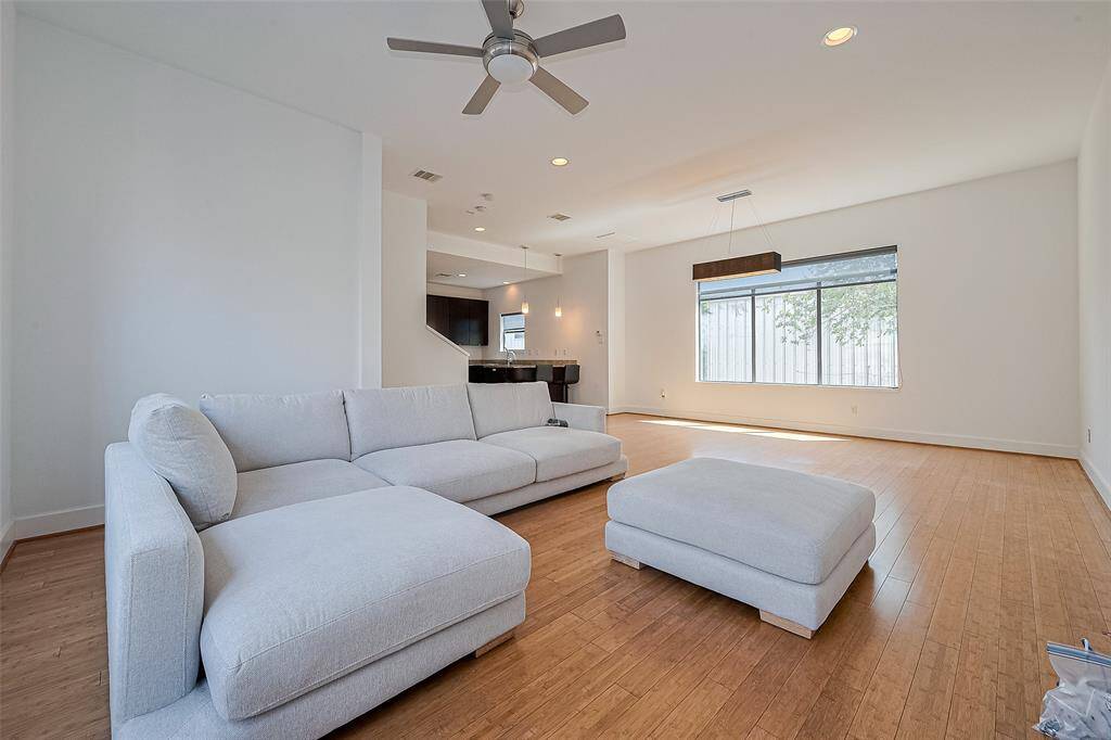 Bright and spacious living room with a modern design, featuring a large sectional sofa and ottoman on light wood flooring. The room is well-lit by a large window and recessed lighting, and includes a ceiling fan for comfort.