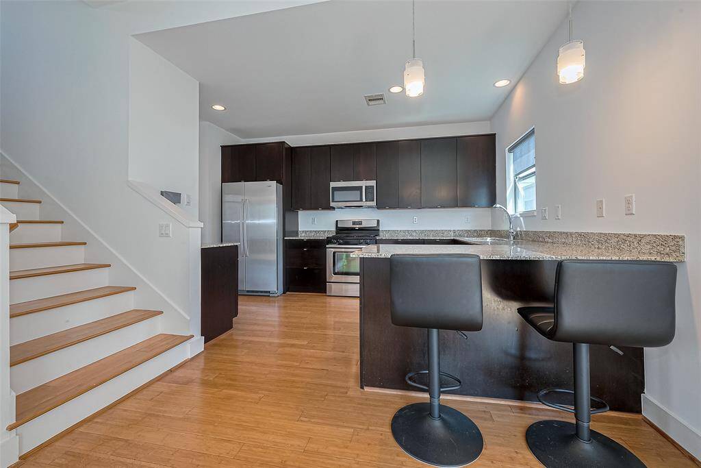 Modern kitchen with dark cabinetry, stainless steel appliances, and a granite countertop. Features an island with bar seating and open layout adjacent to a staircase. Bright and inviting space with ample lighting.