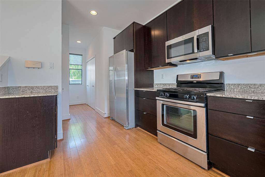 Modern kitchen with sleek dark cabinets, stainless steel appliances, and granite countertops. Features a gas stove, microwave, and large refrigerator. Bright with wood flooring and a window for natural light.