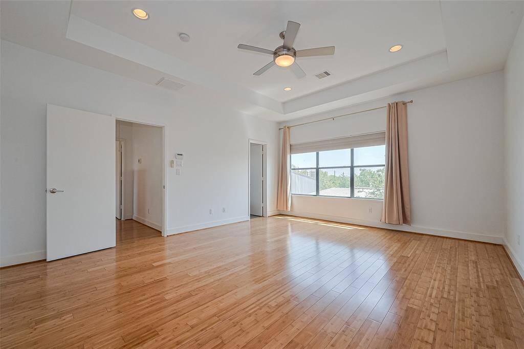 This bright, spacious room features hardwood floors, a ceiling fan, and large windows with curtains, providing ample natural light. The neutral walls and recessed lighting enhance the modern feel, making it a versatile space.