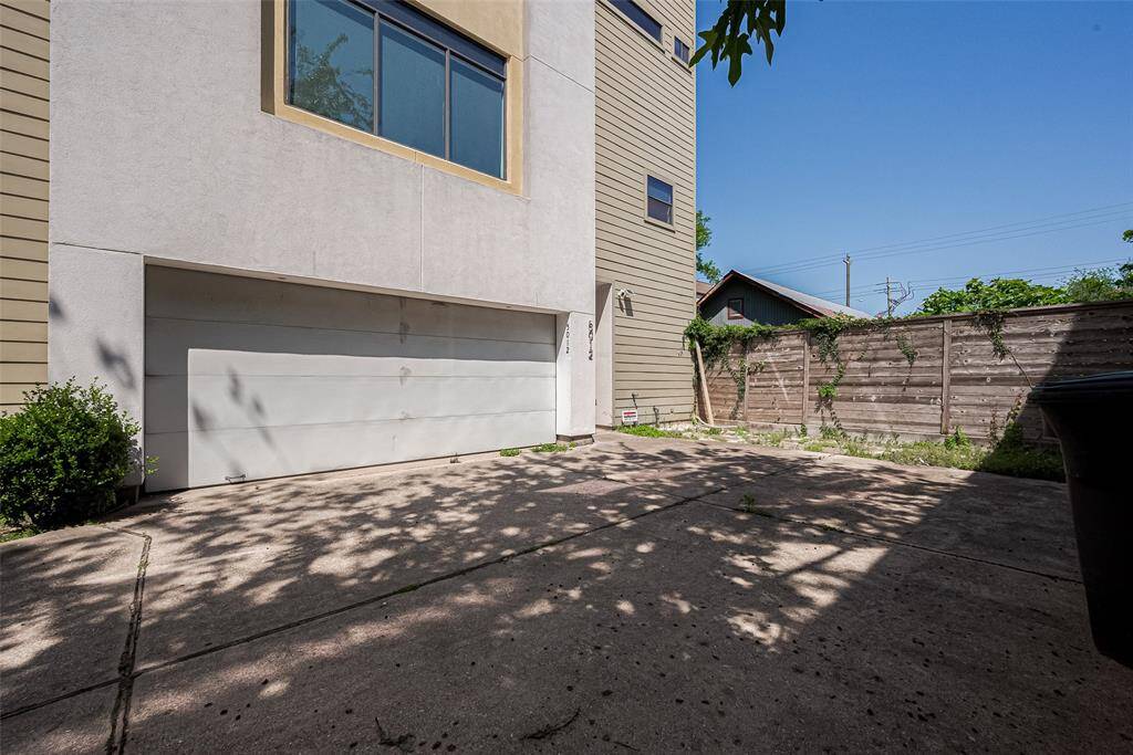 This photo showcases a driveway with a two-car garage attached to a modern home. The area is partially shaded, surrounded by a wooden fence, and features some greenery. The exterior has a mix of light-colored stucco and wood siding.