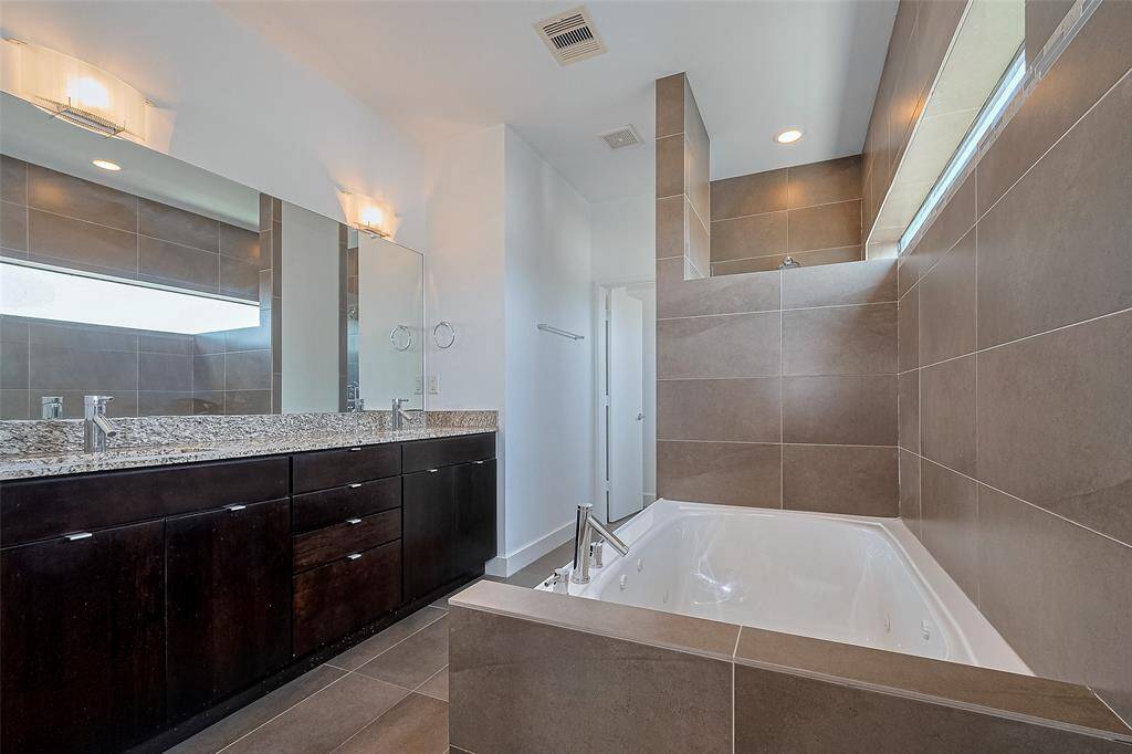 This modern bathroom features a sleek design with a large granite countertop and dual sinks. The spacious soaking tub is surrounded by elegant tile, and there's ample natural light from the high window, creating a relaxing and stylish atmosphere.