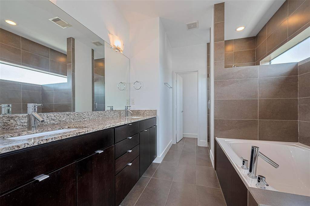This modern bathroom features a sleek double vanity with granite countertops, dark wood cabinets, and a large mirror. The space includes a luxurious soaking tub and is accented with contemporary brown tile, creating a stylish and functional atmosphere.
