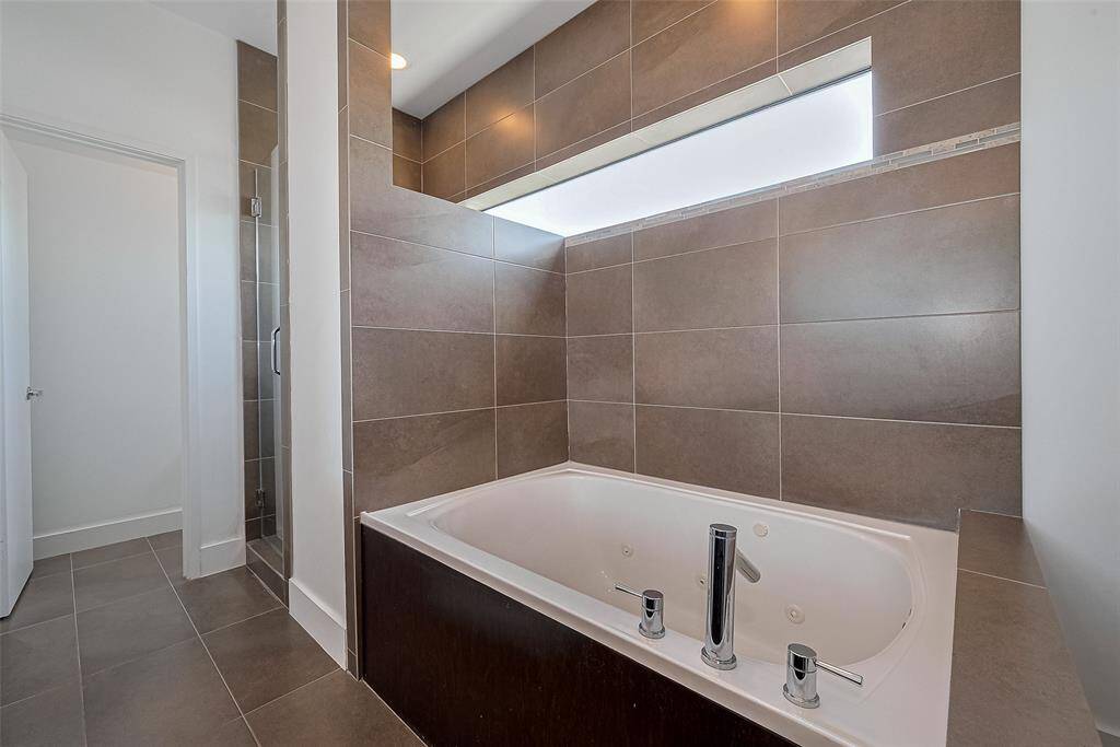 This bathroom features a modern, deep soaking tub with sleek fixtures, surrounded by large, elegant brown tiles. A frosted window above provides natural light while maintaining privacy.