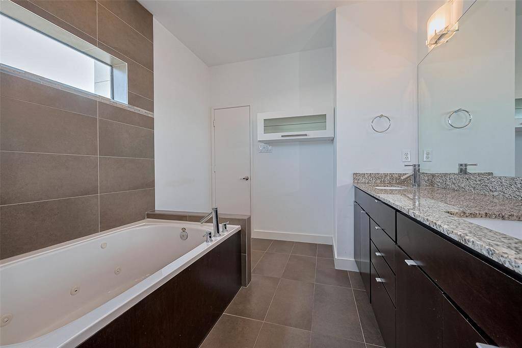 This modern bathroom features a spacious jetted tub, sleek dark wood cabinetry with granite countertops, and a large mirror. The design includes elegant tile flooring and wall accents, creating a clean and contemporary atmosphere.