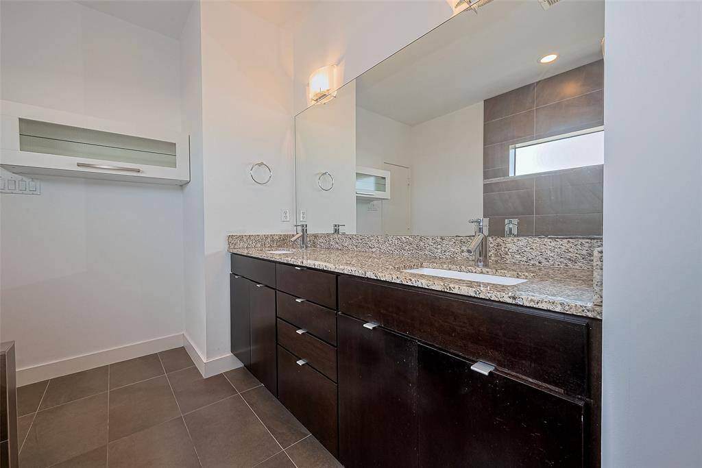 This modern bathroom features a sleek granite countertop with a single sink, dark wood cabinetry, and a large mirror. The space is brightened by recessed lighting and has a minimalistic design with neutral tile flooring.