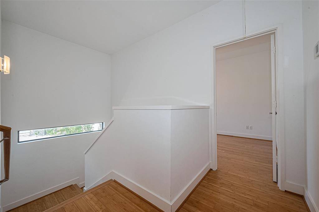 This photo shows a bright, minimalist stairwell with light wood flooring and white walls. A narrow horizontal window allows natural light in, and there's a doorway leading to another room.