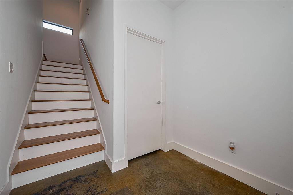 This photo shows a minimalist interior with a staircase featuring wooden steps and a simple handrail. Adjacent is a plain door, and the floor is concrete for a modern touch. The space is well-lit, with a window at the top of the stairs.