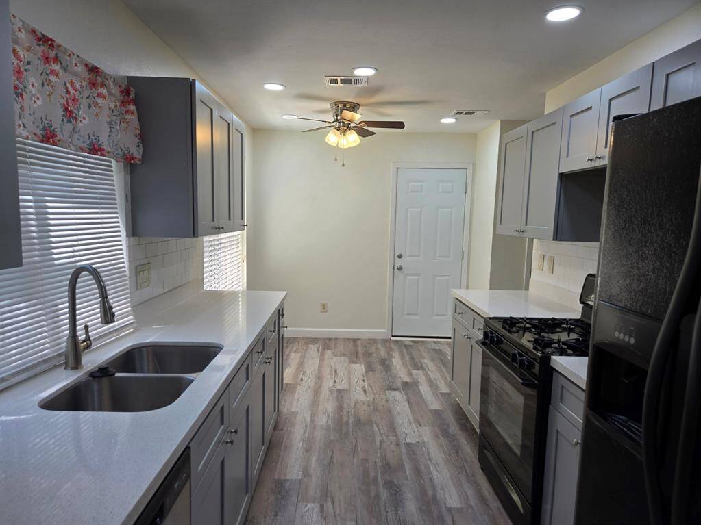 This kitchen features modern gray cabinetry, sleek white countertops, and stainless steel appliances. It has a double sink, wood-look flooring, and ample natural light through the window. A ceiling fan provides additional comfort.