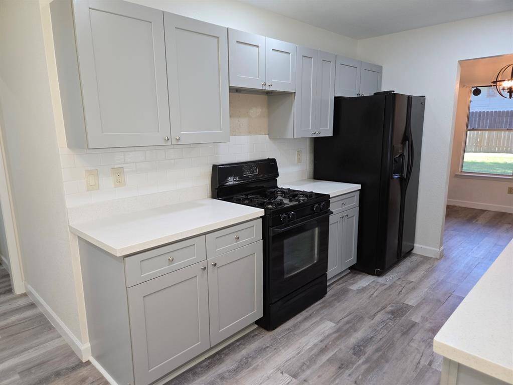 Modern kitchen with light gray cabinets, white countertops, and a black appliance suite including a stove and refrigerator. The space features a subway tile backsplash and laminate wood flooring, offering a sleek and stylish look.