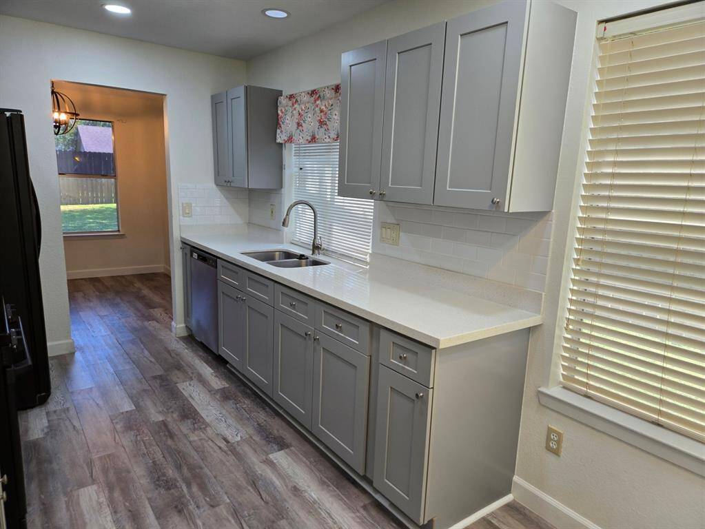 This photo features a modern kitchen with sleek gray cabinets, a white countertop, and a stainless steel dishwasher. The space is well-lit with recessed lighting, and there's a large window with blinds, offering a view of the backyard. The flooring is a wood-look laminate, adding warmth to the room.