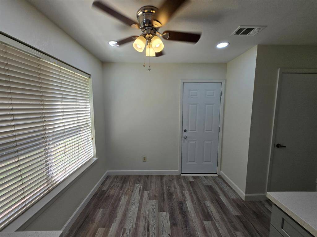 This room features a modern ceiling fan with lights, wood-style flooring, and ample natural light from large windows with blinds. It includes a white door and neutral wall colors, providing a bright and welcoming atmosphere.