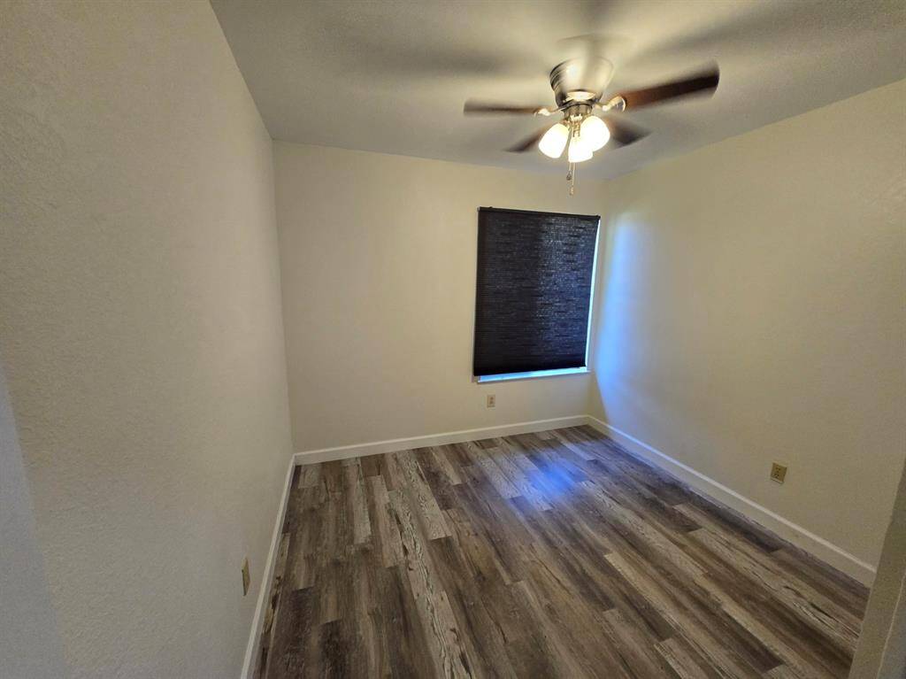 This is a cozy, small room with wood-style flooring, a ceiling fan with lights, and a window with a dark blind, making it suitable for a bedroom or office space.
