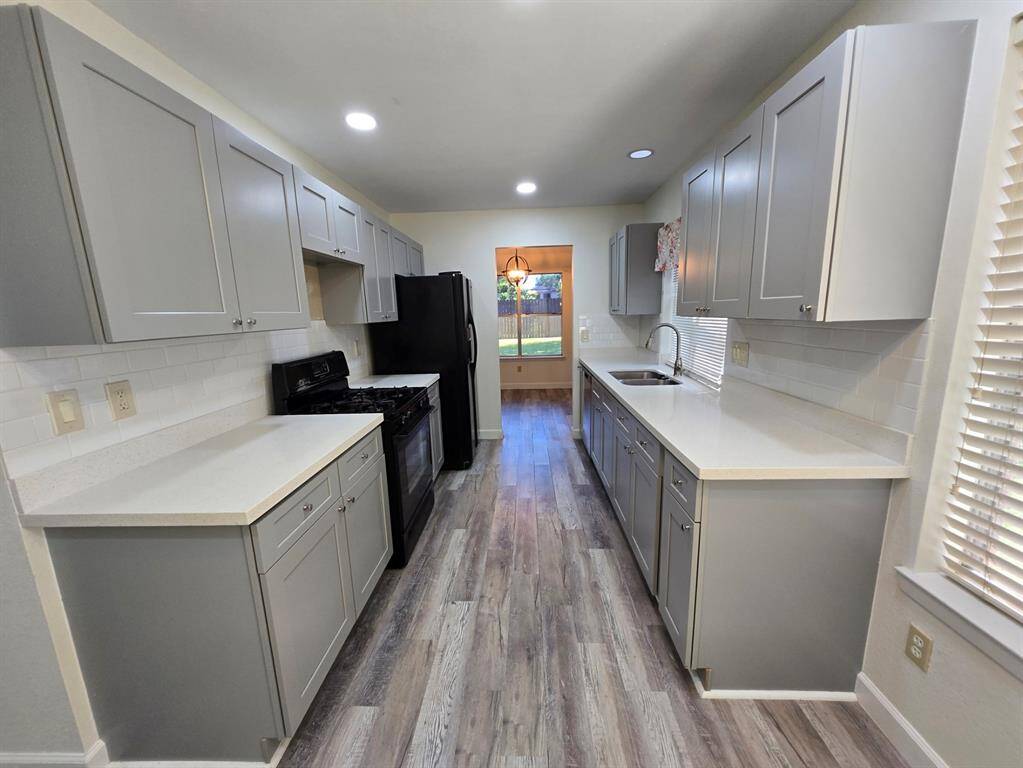 This kitchen features modern gray cabinetry, sleek white countertops, and stainless steel appliances. The wood-look flooring adds warmth, and ample natural light comes through the window, offering a pleasant view of the backyard.