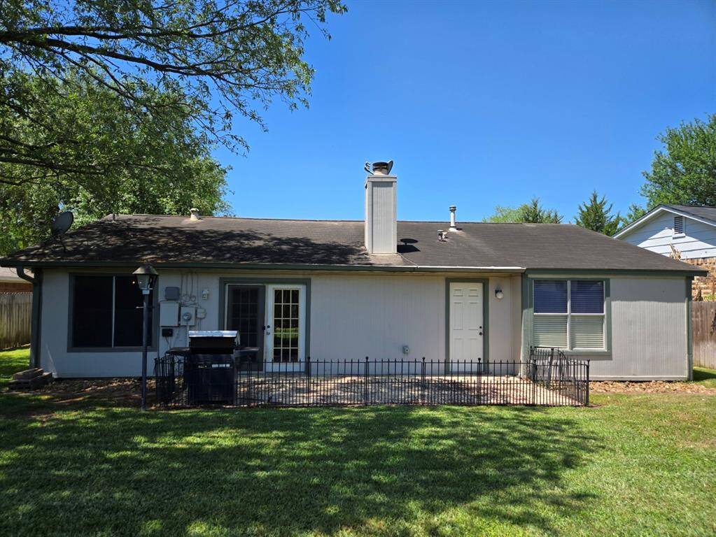 This photo shows the back of a single-story home with a simple, neat exterior. It features a small patio area, a chimney, and a fenced-in section. The yard is grassy with mature trees providing shade.