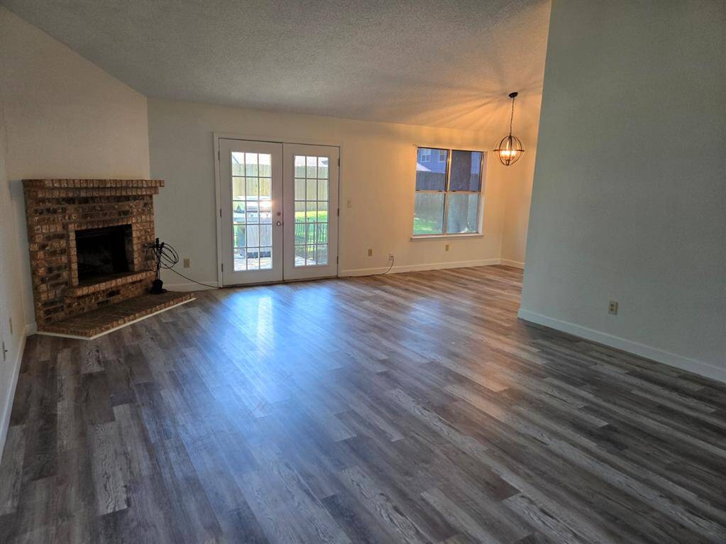 This spacious living room features sleek wood flooring, a cozy brick fireplace, and ample natural light from large windows and French doors leading to the backyard. A modern light fixture adds a touch of elegance.