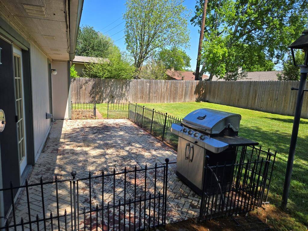 This backyard features a spacious patio with a built-in grill, surrounded by a small fence, perfect for outdoor entertaining. It opens to a grassy area with mature trees and a wooden privacy fence.