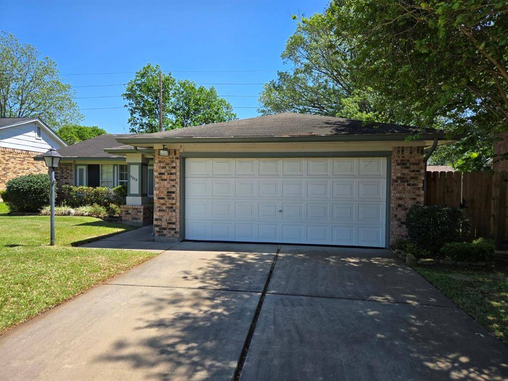 This photo shows a charming single-story brick home with a two-car garage and a well-maintained front yard. The driveway is spacious, and there are trees providing some shade. The house has a welcoming and cozy curb appeal.