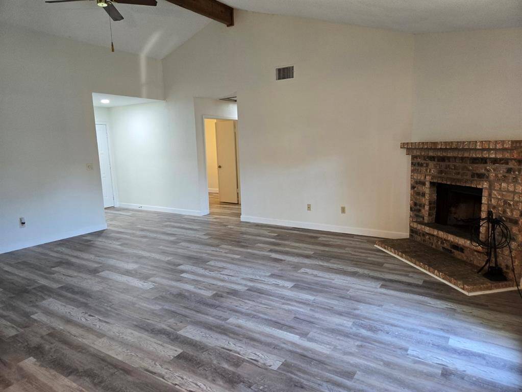 This spacious living room features light wood flooring, a brick fireplace, and high ceilings with exposed beams, offering a cozy yet open atmosphere.