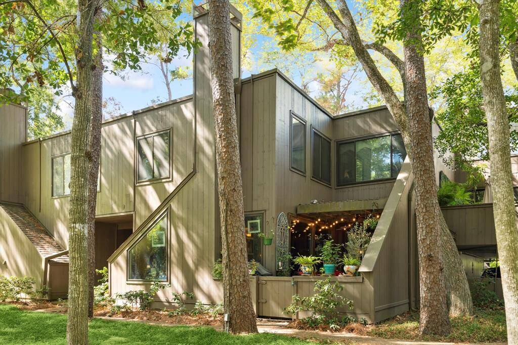 This is a two-story contemporary home featuring a neutral color palette, nestled among mature trees that offer ample shade and privacy. The architecture includes interesting angles and large windows, with a cozy entrance and a welcoming, covered outdoor space on the side.