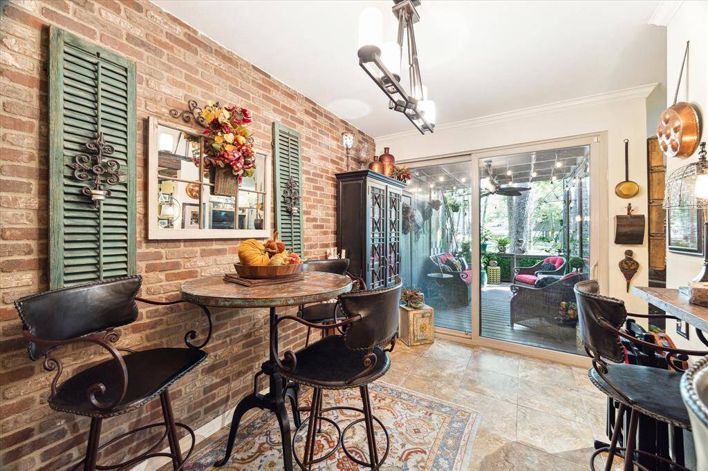 This is a cozy, rustic-style kitchen nook featuring exposed brick walls, a wood shelving unit filled with various decorative items, and a small round table with iron-based stools. The space is accented with colorful décor and green window shutters.