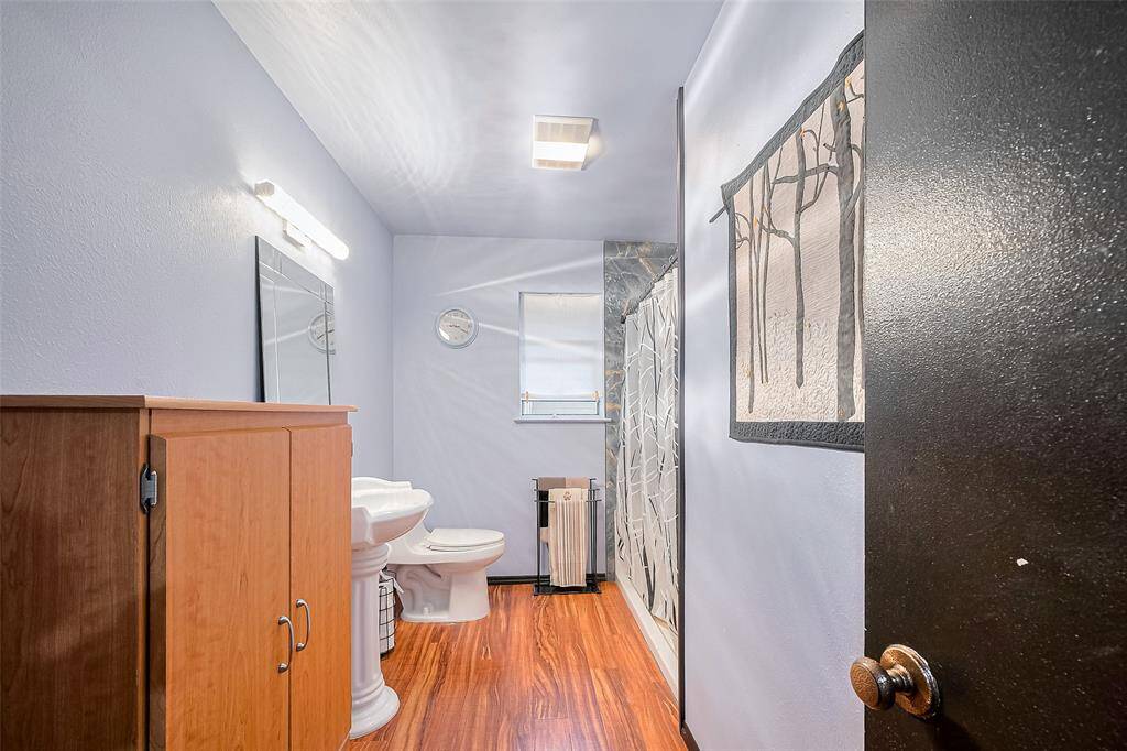 The second full bathroom features a clean, modern aesthetic with cool-toned walls, beautiful lighting, and rich wood-look flooring. A pedestal sink, walk-in shower, and natural light from the window make this space feel fresh and functional—ideal for guests or shared use.