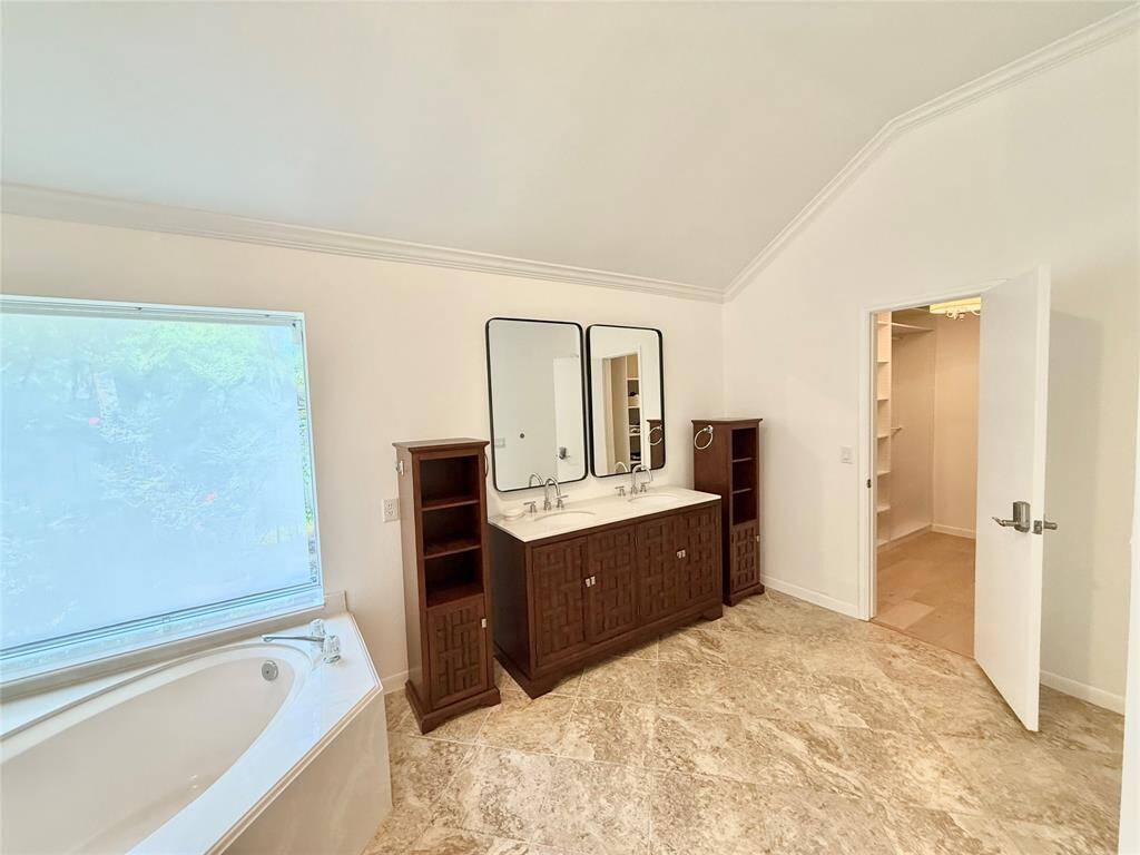 This spacious bathroom features a luxurious soaking tub, a double vanity with two mirrors, elegant wood cabinetry, and a large window providing natural light. Adjacent is a walk-in closet with ample shelving.