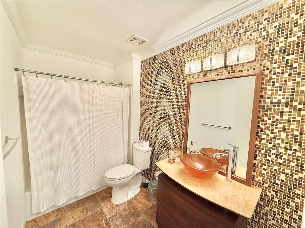 This bathroom features a stylish mosaic tile accent wall, modern vanity with a glass vessel sink, and a shower-tub combo. The space is well-lit with elegant lighting fixtures and has a cozy, contemporary feel.